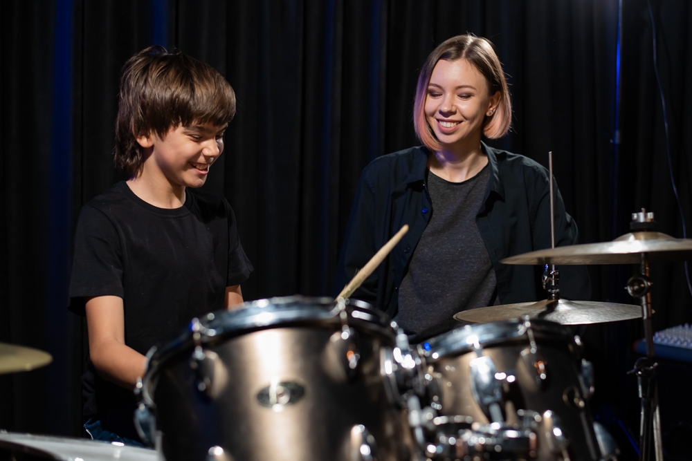 Man kan spille trommer som gutten på bilde både i korps og i band. Vi ser også en lærerinne som underviser gutten på bildet. 